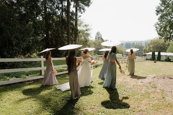 A bride and bridesmaids in long dresses holding parasols and bouquets, walking on a grassy lawn near a white fence.