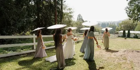 A bride and bridesmaids in long dresses holding parasols and bouquets, walking on a grassy lawn near a white fence.