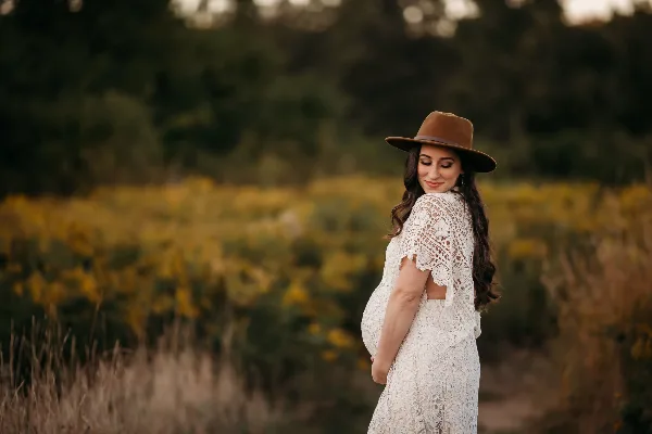 Professional outdoor maternity portrait of a pregnant woman in a white lace dress and brown hat standing in a field of yellow flowers.