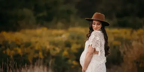Professional outdoor maternity portrait of a pregnant woman in a white lace dress and brown hat standing in a field of yellow flowers.