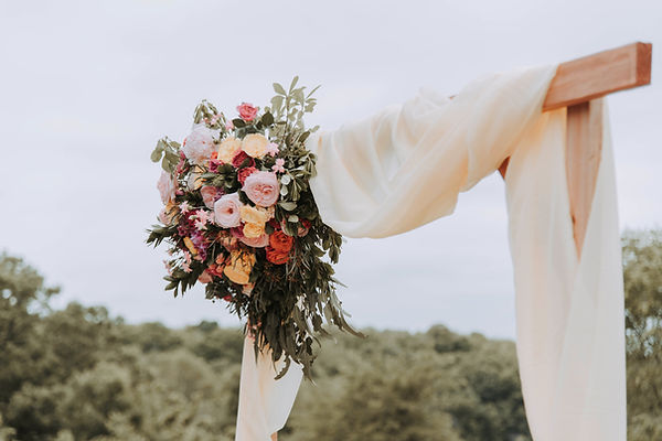 A wooden wedding arch decorated with a large bouquet of pink and orange flowers and white fabric draping, set against an outdoor background.