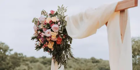 A wooden wedding arch decorated with a large bouquet of pink and orange flowers and white fabric draping, set against an outdoor background.