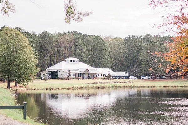 A white event venue building with a cupola situated on a grassy lawn next to a private lake, surrounded by a dense forest.