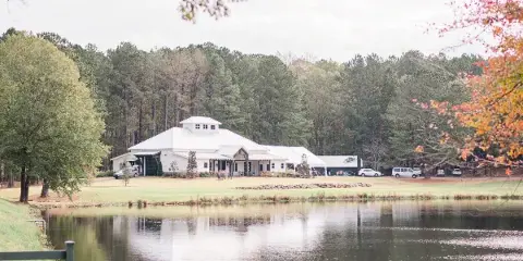 A white event venue building with a cupola situated on a grassy lawn next to a private lake, surrounded by a dense forest.