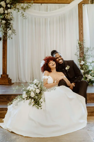 A bride and groom laughing together on a wooden platform in front of a white draped archway with floral decorations.