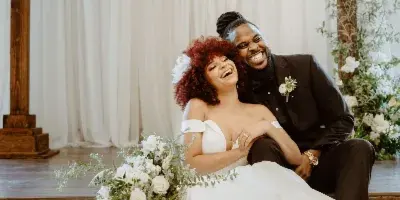 A bride and groom laughing together on a wooden platform in front of a white draped archway with floral decorations.