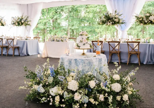 A wedding reception table setting featuring a white wedding cake, blue and white floral arrangements, and cross-back chairs under a white tent.