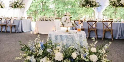 A wedding reception table setting featuring a white wedding cake, blue and white floral arrangements, and cross-back chairs under a white tent.