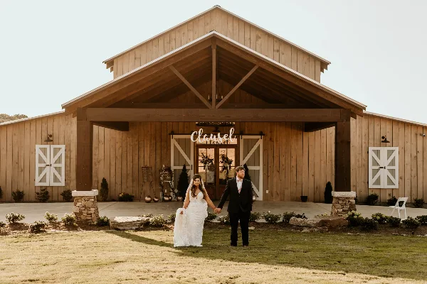A bride and groom standing in front of a large wooden barn wedding venue with a 'Classel' sign above the entrance.