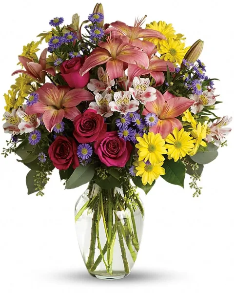 A vibrant floral arrangement featuring pink lilies, red roses, yellow daisies, and purple asters in a clear glass vase.