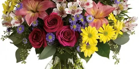 A vibrant floral arrangement featuring pink lilies, red roses, yellow daisies, and purple asters in a clear glass vase.