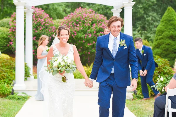 A bride in a white lace gown and a groom in a blue suit walking hand-in-hand down a wedding aisle under a white arbor.