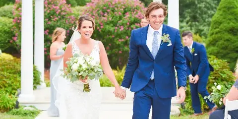 A bride in a white lace gown and a groom in a blue suit walking hand-in-hand down a wedding aisle under a white arbor.