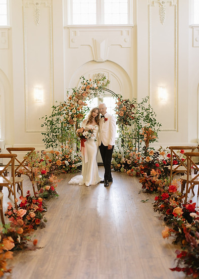 A bride and groom standing in a wedding venue aisle decorated with a large floral arch and flowers lining the walkway.