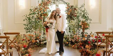 A bride and groom standing in a wedding venue aisle decorated with a large floral arch and flowers lining the walkway.