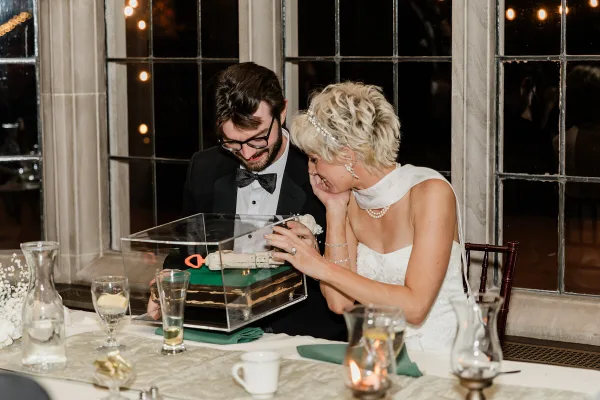 A bride and groom smiling together at a wedding reception table, looking at a cake in a clear box.