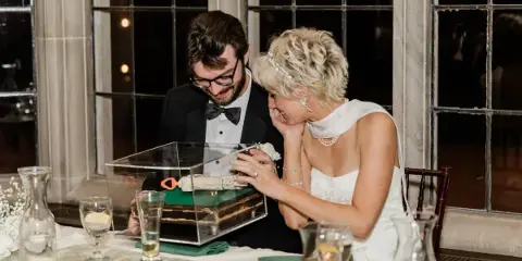 A bride and groom smiling together at a wedding reception table, looking at a cake in a clear box.