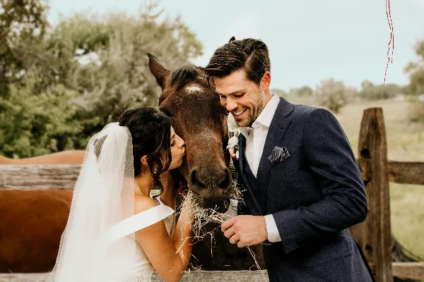 A bride and groom smiling while feeding a horse at a rustic outdoor wedding venue.