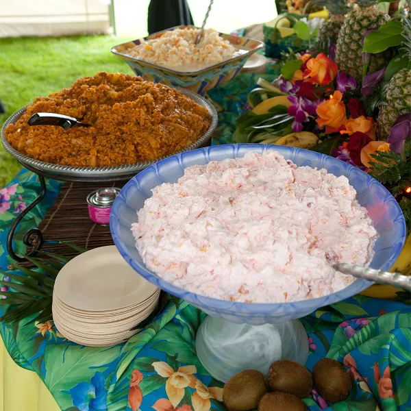 Outdoor catering buffet table with large bowls of prepared food, serving utensils, plates, and floral decorations.
