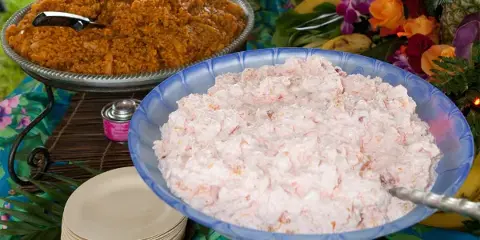 Outdoor catering buffet table with large bowls of prepared food, serving utensils, plates, and floral decorations.