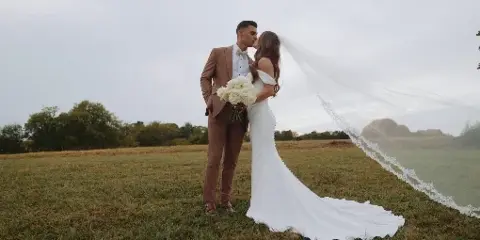 A bride and groom sharing a kiss in a grassy field, with the bride holding a bouquet of white roses.