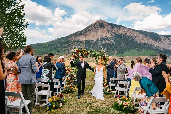 A bride and groom walking down the aisle at an outdoor wedding ceremony with a mountain in the background.