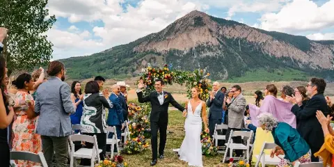 A bride and groom walking down the aisle at an outdoor wedding ceremony with a mountain in the background.