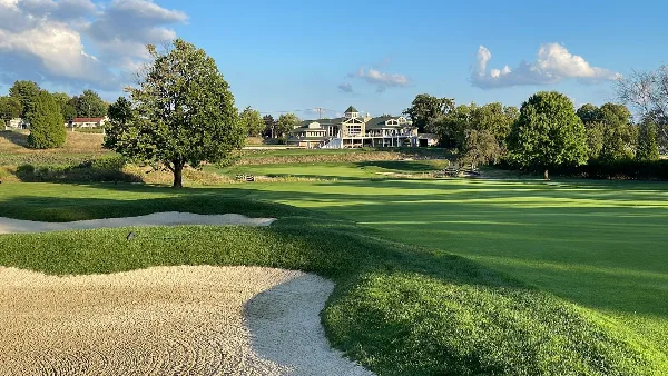 A wide shot of a green golf course with sand bunkers in the foreground and a large clubhouse building in the background under a blue sky.