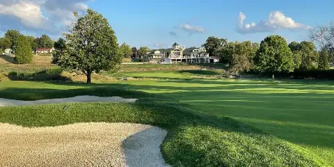 A wide shot of a green golf course with sand bunkers in the foreground and a large clubhouse building in the background under a blue sky.