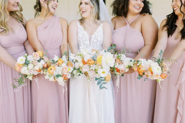 A bride and bridesmaids in pink dresses holding a long, cascading wedding bouquet with white and peach flowers.