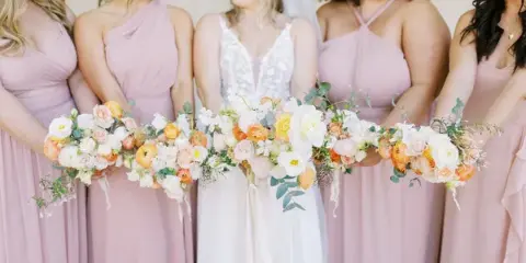 A bride and bridesmaids in pink dresses holding a long, cascading wedding bouquet with white and peach flowers.