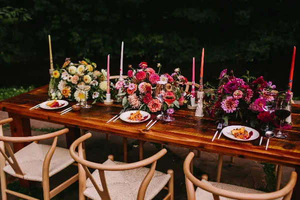A rustic wooden dining table set outdoors with vibrant floral centerpieces, tall candles, and place settings.
