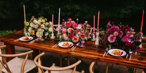 A rustic wooden dining table set outdoors with vibrant floral centerpieces, tall candles, and place settings.