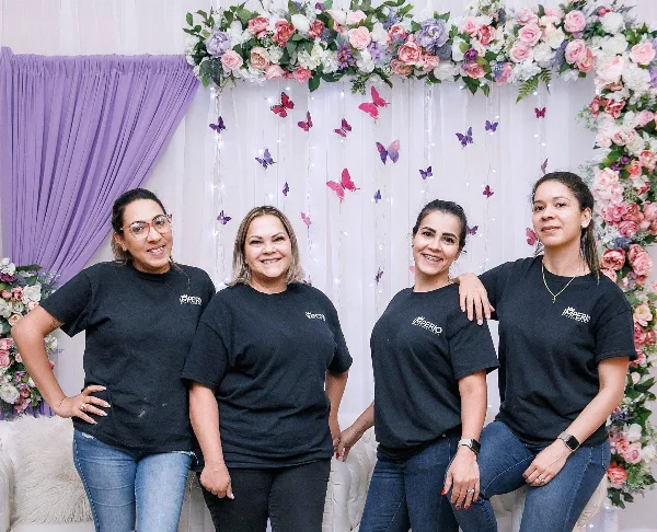Four women in black Imperio Decor & Event Rental shirts posing in front of a decorated event backdrop with flowers, draping, and butterfly accents.