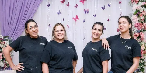 Four women in black Imperio Decor & Event Rental shirts posing in front of a decorated event backdrop with flowers, draping, and butterfly accents.
