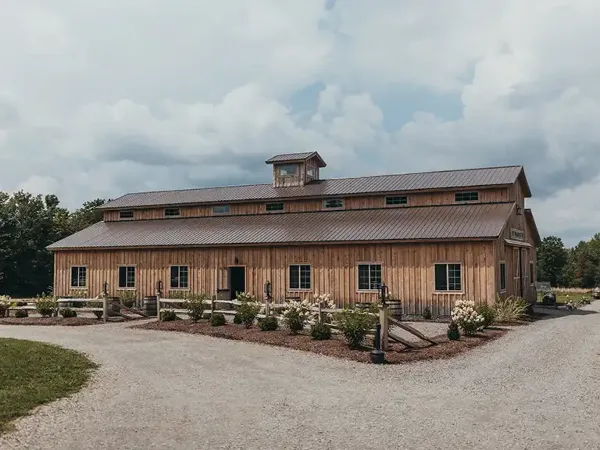 Exterior view of a large rustic wooden barn with a metal roof and cupola, surrounded by a gravel driveway and landscaped flower beds.