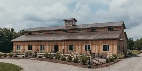 Exterior view of a large rustic wooden barn with a metal roof and cupola, surrounded by a gravel driveway and landscaped flower beds.