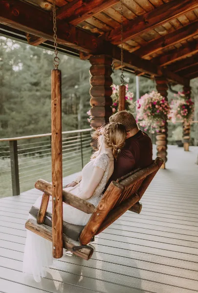 A bride and groom sitting together on a wooden swing on a rustic porch, with the bride in a white lace dress and the groom in a dark shirt.