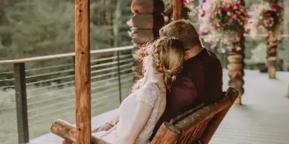 A bride and groom sitting together on a wooden swing on a rustic porch, with the bride in a white lace dress and the groom in a dark shirt.