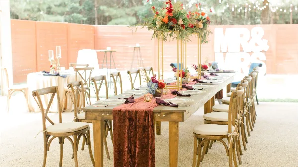 A long wooden banquet table set with a rust-colored runner, floral arrangements, and place settings in an outdoor venue.