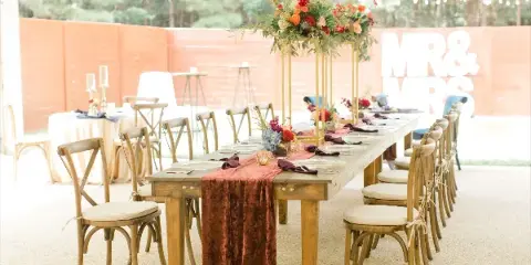 A long wooden banquet table set with a rust-colored runner, floral arrangements, and place settings in an outdoor venue.