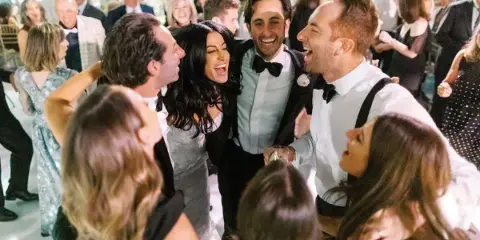 A group of wedding guests dancing and laughing on a dance floor at a reception.