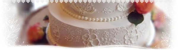 A close-up photograph of a multi-tiered white wedding cake decorated with intricate floral piping, pearl trim, and fresh orange roses.