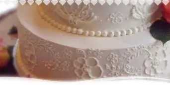 A close-up photograph of a multi-tiered white wedding cake decorated with intricate floral piping, pearl trim, and fresh orange roses.