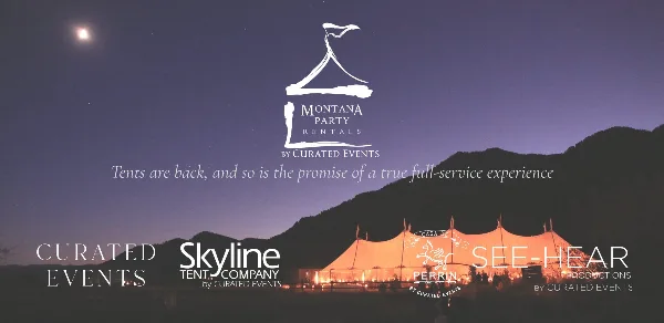 A large, illuminated event tent set up in a field at night with mountains in the background.