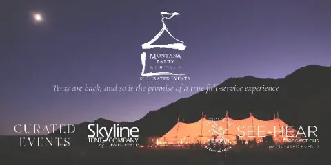 A large, illuminated event tent set up in a field at night with mountains in the background.