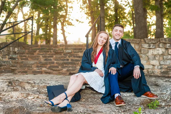 A professional portrait of a smiling couple wearing graduation gowns and caps, sitting on stone steps outdoors.