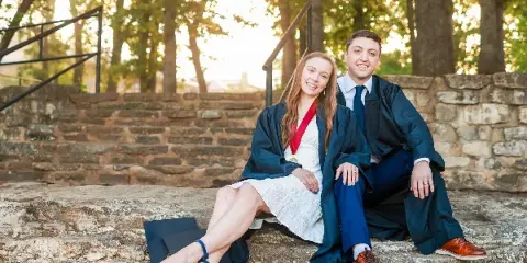 A professional portrait of a smiling couple wearing graduation gowns and caps, sitting on stone steps outdoors.