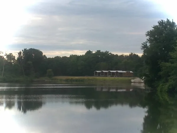 A wide shot of a calm lake with a long wooden barn structure and trees on the far shore.