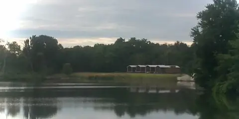 A wide shot of a calm lake with a long wooden barn structure and trees on the far shore.